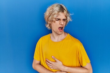 Young modern man standing over blue background with hand on stomach because indigestion, painful illness feeling unwell. ache concept.