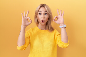 Young caucasian woman wearing yellow sweater looking surprised and shocked doing ok approval symbol with fingers. crazy expression