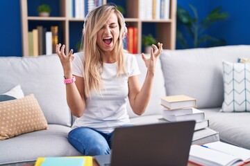 Young blonde woman studying using computer laptop at home crazy and mad shouting and yelling with aggressive expression and arms raised. frustration concept.