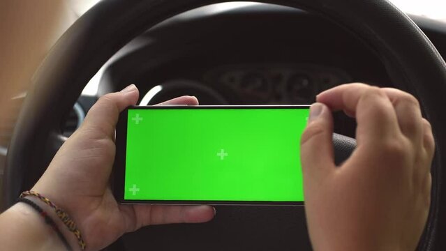Close up of a woman's hands holding a mobile telephone with a vertical green screen in car. Woman's hands zoom in on the screen with two fingers on the background of the steering wheel. Chroma key, sm