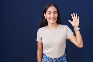 Young hispanic woman standing over blue background showing and pointing up with fingers number five while smiling confident and happy.