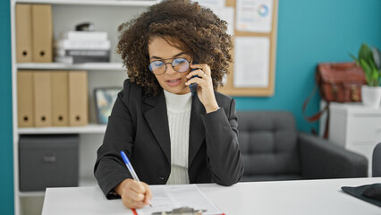 Obraz premium Young beautiful hispanic woman business worker talking on smartphone writing on document at the office