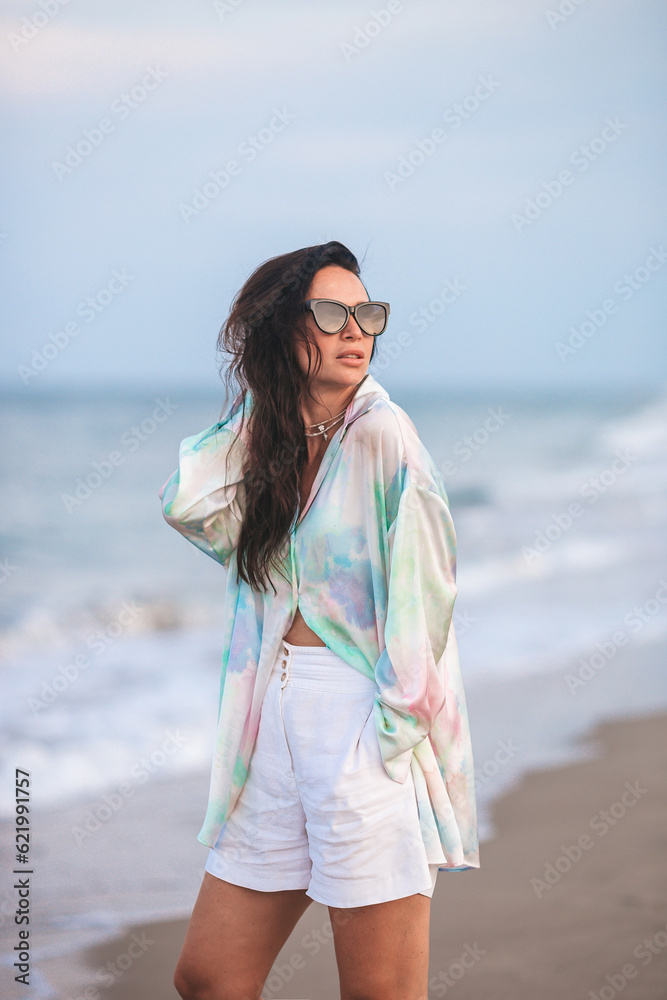 Wall mural Young happy woman on the beach enjoy her summer vacation. Woman walking along the sea in evening. 