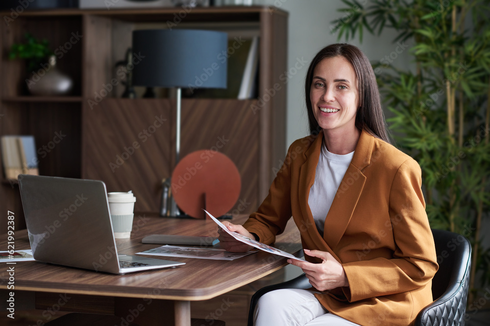 Aufkleber young smiling female designer with paperwork, looking at camera with smile while sitting by workplac
