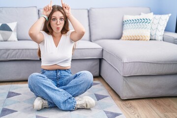 Young caucasian woman sitting on the floor at the living room doing funny gesture with finger over head as bull horns