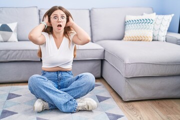 Obraz premium Young caucasian woman sitting on the floor at the living room crazy and scared with hands on head, afraid and surprised of shock with open mouth