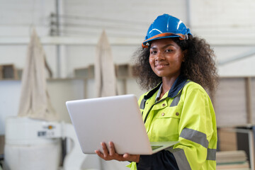 African American woman engineer with laptop computer working in furniture factory. Female technical wearing vest and helmet safety standing workshop making wooden product.