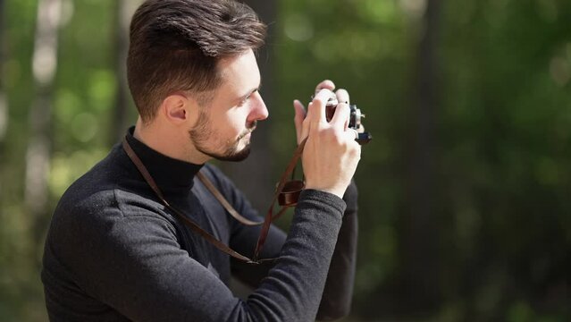 Side view portrait of smiling Caucasian young man taking photos of nature with camera standing in sunshine in summer forest. Happy confident tourist enjoying leisure on vacations