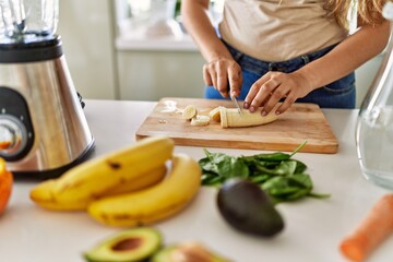 Young beautiful hispanic woman preparing vegetable smoothie with blender cutting banana at the kitchen