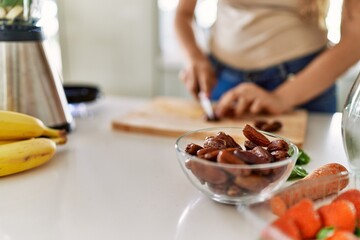 Young beautiful hispanic woman preparing vegetable smoothie with blender cutting date at the kitchen
