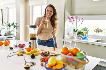 Young beautiful hispanic woman smiling confident holding glass of vegetable smoothie at the kitchen