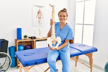 Young beautiful hispanic woman physiotherapist smiling confident holding anatomical model of spinal column at rehab clinic - obrazy, fototapety, plakaty