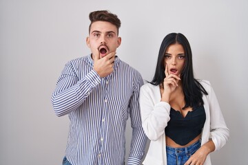 Young hispanic couple standing over white background looking fascinated with disbelief, surprise and amazed expression with hands on chin