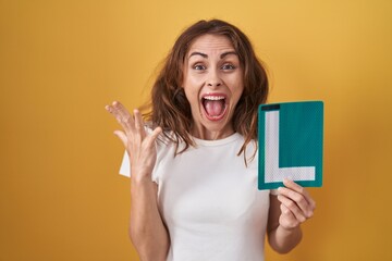 Beautiful brunette woman holding l sign for new driver celebrating victory with happy smile and winner expression with raised hands