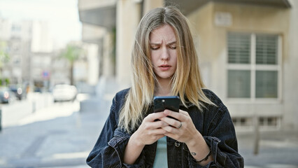 Young blonde woman using smartphone with serious face at street