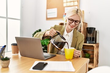 Young blonde woman business worker using laptop pouring coffee on cup at office