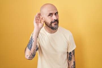 Hispanic man with tattoos standing over yellow background smiling with hand over ear listening an hearing to rumor or gossip. deafness concept.