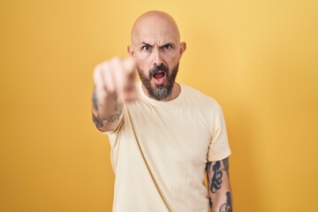 Hispanic man with tattoos standing over yellow background pointing displeased and frustrated to the...