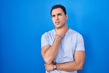 Young hispanic man standing over blue background touching painful neck, sore throat for flu, clod and infection