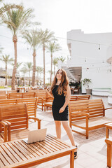 A woman remotely works on the cafe terrace with a laptop and phone in a country where it is always summer. A girl with an online profession works from anywhere in the world, travels in a warm climate