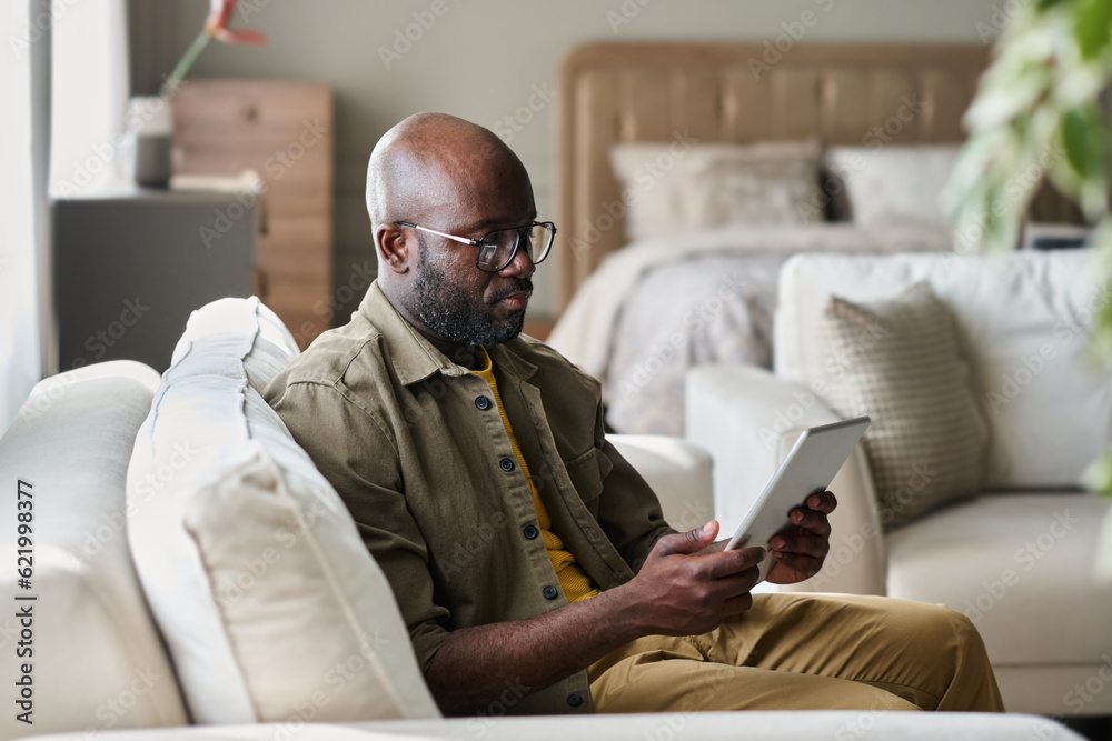 Aufkleber Young serious businessman looking at screen of tablet while sitting on soft grey comfortable couch in living room and scrolling through data