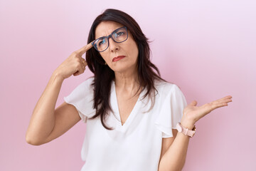 Middle age hispanic woman wearing casual white t shirt and glasses confused and annoyed with open palm showing copy space and pointing finger to forehead. think about it.