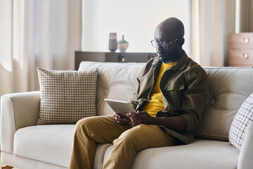 Young African American male freelancer with tablet sitting on soft comfortable couch in living room and looking through online data