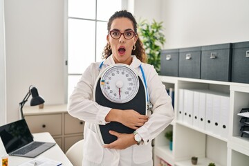 Young hispanic woman working at dietitian clinic afraid and shocked with surprise and amazed...