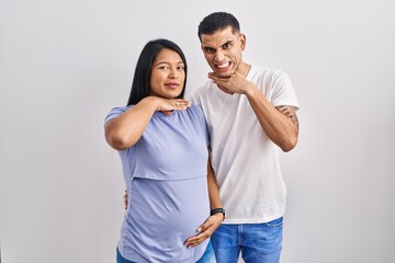 Young hispanic couple expecting a baby standing over background cutting throat with hand as knife,...