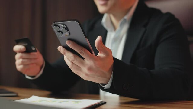 Close up of black smartphone and stylish businessman entering credit card data for making online payment on website at work. Successful asian executive using modern remote way of bank transactions.
