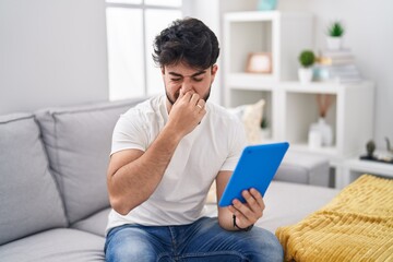 Hispanic man with beard using touchpad sitting on the sofa smelling something stinky and disgusting, intolerable smell, holding breath with fingers on nose. bad smell - obrazy, fototapety, plakaty