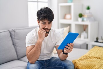 Hispanic man with beard using touchpad sitting on the sofa mouth and lips shut as zip with fingers....