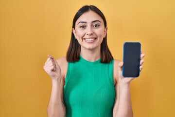 Hispanic girl holding smartphone showing screen screaming proud, celebrating victory and success...
