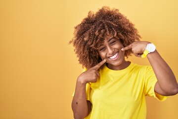 Young hispanic woman with curly hair standing over yellow background smiling cheerful showing and pointing with fingers teeth and mouth. dental health concept.