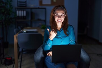 Brunette woman working at the office at night smiling with happy face looking and pointing to the side with thumb up.