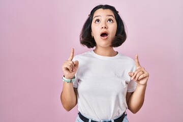 Young hispanic woman wearing casual white t shirt over pink background amazed and surprised looking up and pointing with fingers and raised arms.