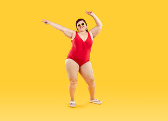 Fototapeta na wymiar Full length happy smiling fat woman wearing red plus size swimsuit, beach flip flops, sunglasses and earrings dancing on yellow background, doing peace love victory hand sign gesture. Swimwear concept