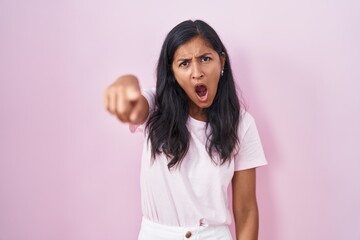 Young hispanic woman standing over pink background pointing displeased and frustrated to the...