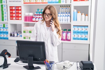 Young woman pharmacist talking on smartphone using computer at pharmacy