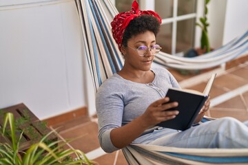 African american woman reading book lying on hammock at home terrace