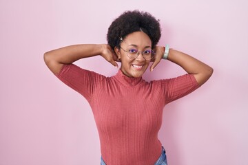 Beautiful african woman with curly hair standing over pink background relaxing and stretching, arms and hands behind head and neck smiling happy