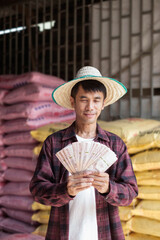 Obraz premium Asian farmer man wear red shirt standing and holding Thai banknote with chemical fertilizer sack at warehouse