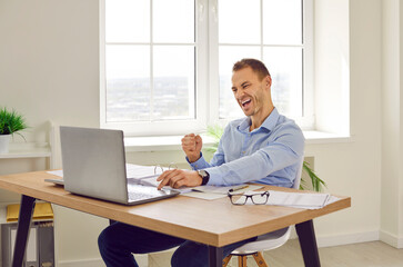 Fototapeta premium Happy business man celebrating success in office. Young man sitting at working desk, looking at laptop computer, doing fist gesture, and saying Yes, I did it. Success, victory, satisfaction concept