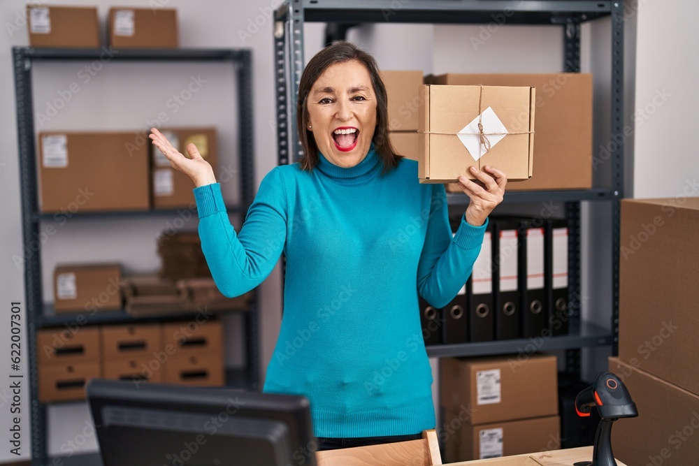 Wall mural Middle age hispanic woman working at small business ecommerce holding parcel celebrating achievement with happy smile and winner expression with raised hand