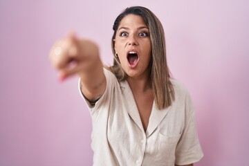 Blonde woman standing over pink background pointing with finger surprised ahead, open mouth amazed expression, something on the front
