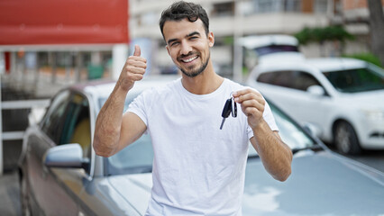 Young hispanic man holding key of new car doing thumb up gesture at street