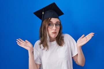 Blonde caucasian woman wearing graduation cap clueless and confused expression with arms and hands...