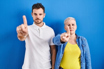 Young brazilian mother and son standing over blue background pointing with finger up and angry expression, showing no gesture