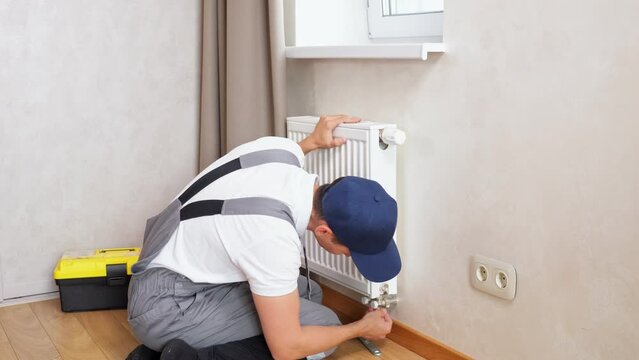 Repairman repairs heating battery with temperature control in room of apartment by turning bolt with wrench. Process by employee. Home service concept