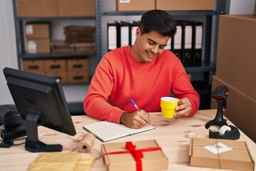 Young hispanic man ecommerce business worker writing on notebook drinking coffee at office